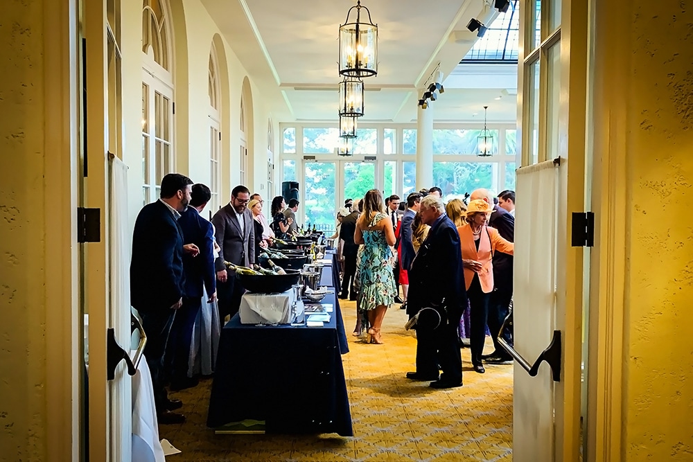 Crowd of guests mingling at a fundraiser event inside a well-lit venue, with tables displaying items for auction and a festive atmosphere.