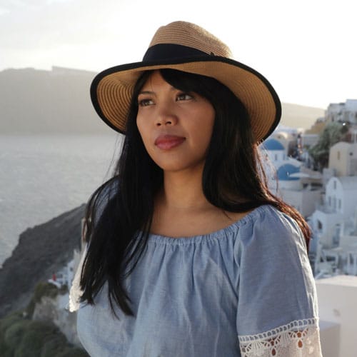 Woman with long black hair wearing a straw hat and blue off-shoulder top, standing by the seaside with white buildings in the background, highlighting a relaxed and elegant look suitable for wedding hair inspiration.