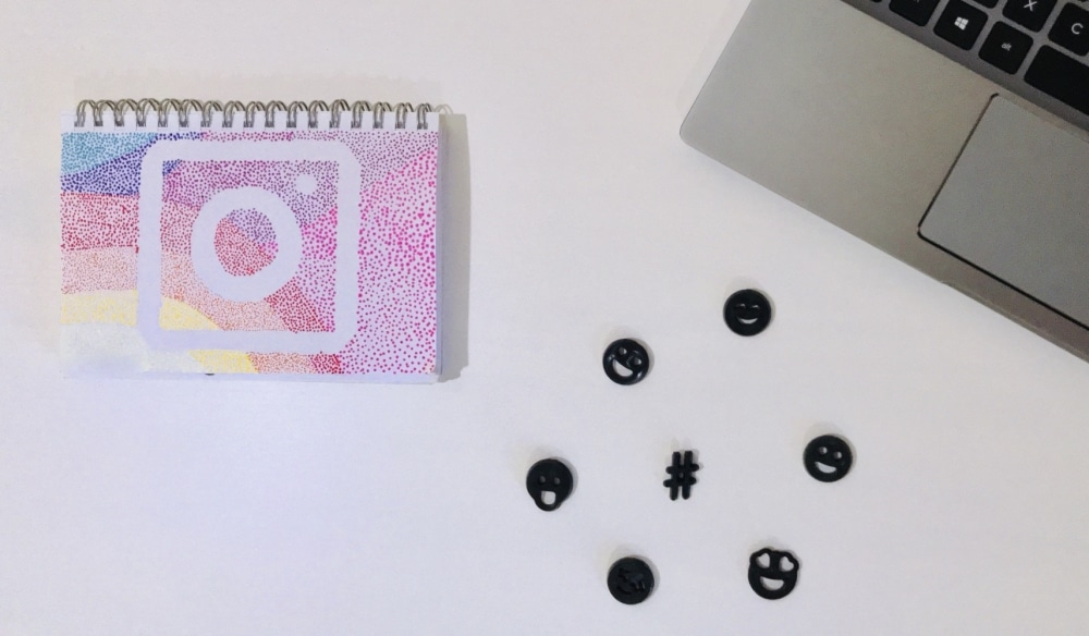 Colorful notebook featuring Instagram logo, surrounded by black emoji and hashtag symbols, next to a laptop, symbolizing social media engagement and event promotion strategies.