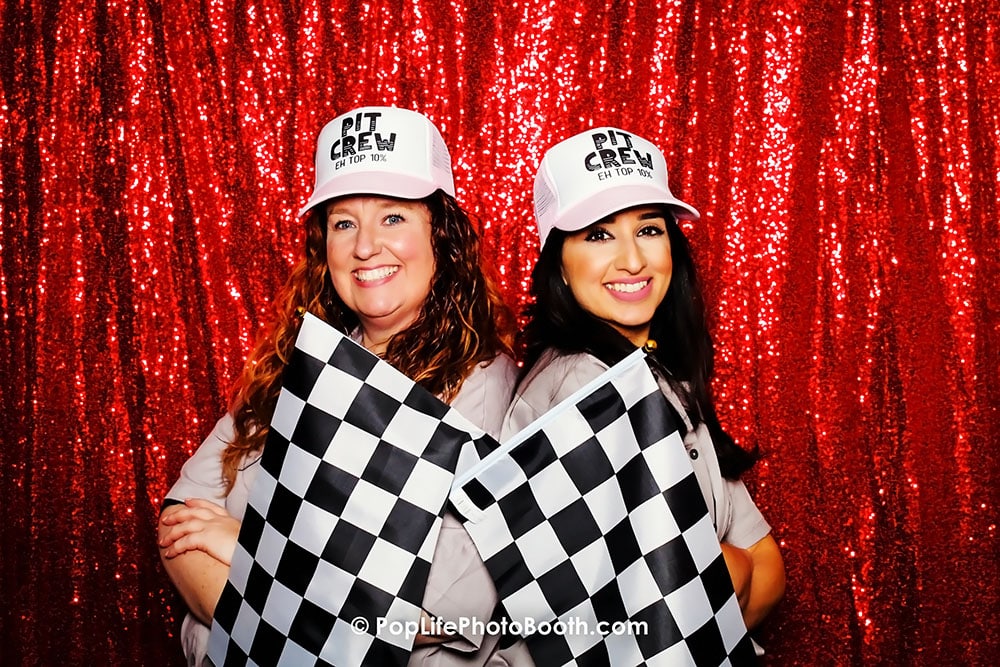 Two women wearing "Pit Crew" hats holding checkered flags in front of a sparkling red backdrop, showcasing a fun and festive atmosphere for corporate events.
