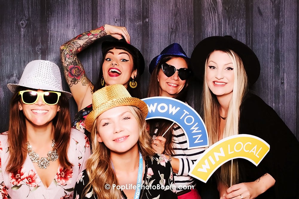 Group of five women in stylish hats and sunglasses posing in a photo booth, holding signs that read "WOW TOWN" and "WIN LOCAL," showcasing a fun atmosphere at a corporate event.