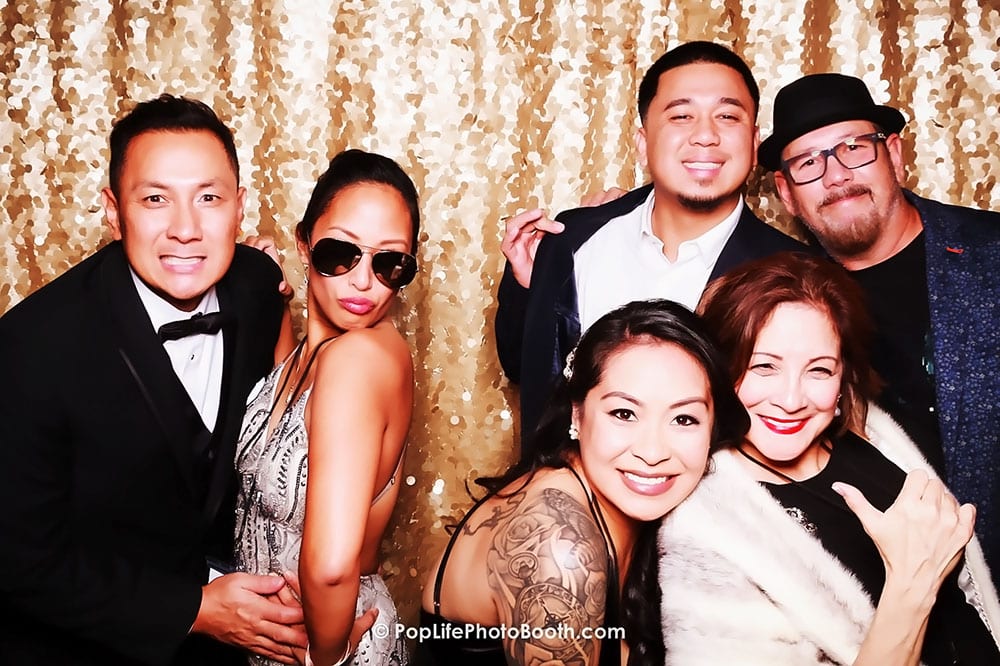 Group of stylishly dressed corporate guests posing playfully in a photo booth, with a shimmering gold backdrop, showcasing vibrant expressions and camaraderie.