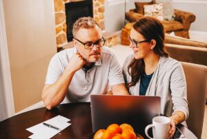 Couple planning holiday event at home, discussing details over a laptop, with coffee and oranges on the table.