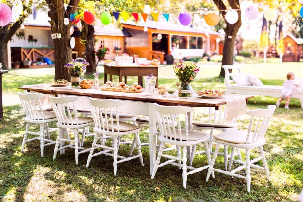 Garden party table set with white chairs, vibrant floral arrangements, and assorted food displays, surrounded by festive decorations and a sunny outdoor setting, ideal for summer weddings and celebrations.