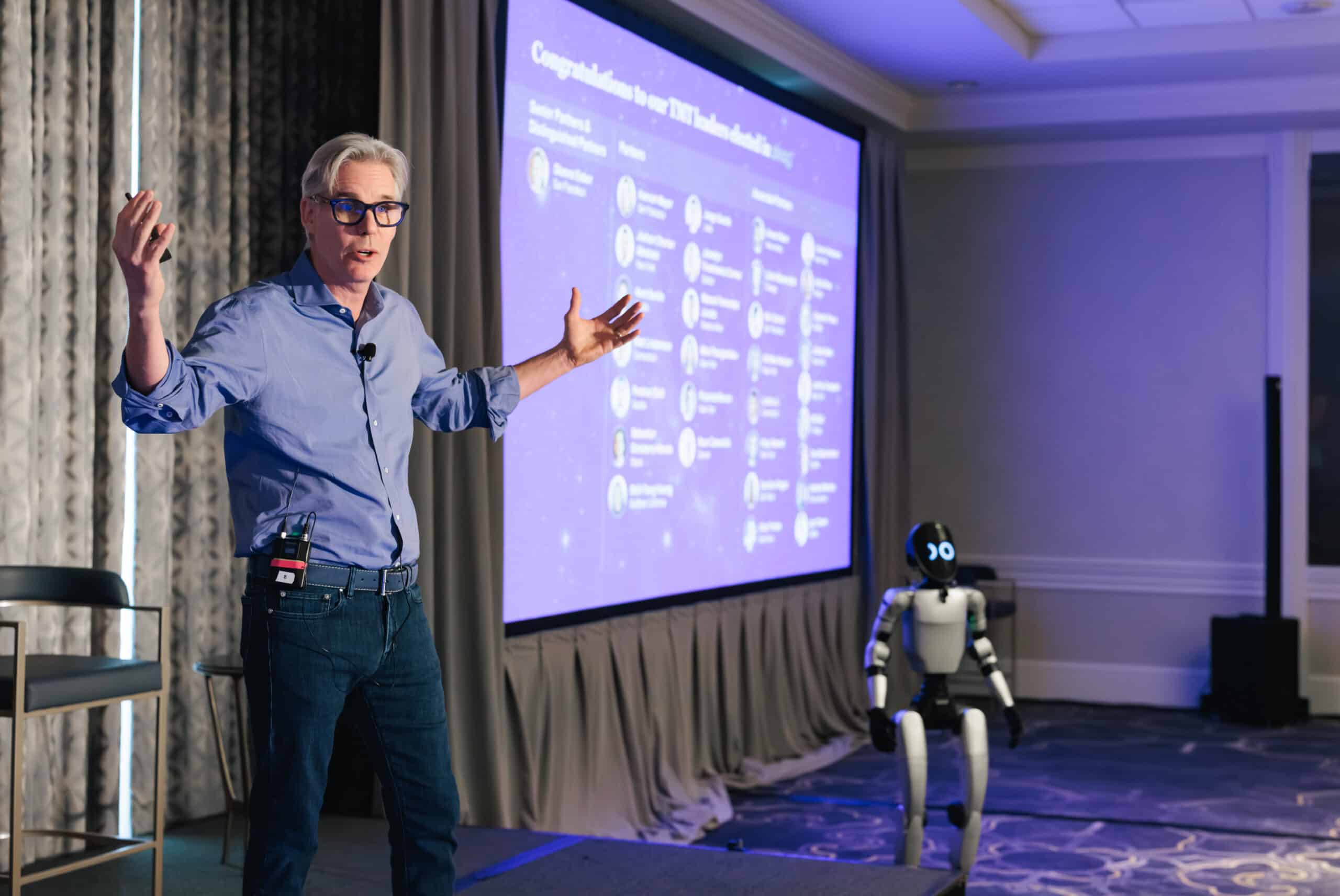 Speaker presenting at event with screen displaying data and a robot on stage.
