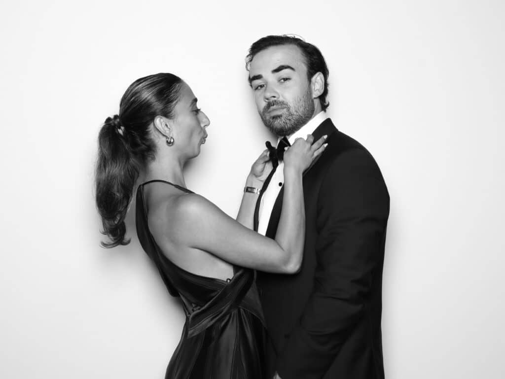 Couple in formal attire sharing a playful, intimate moment, with the woman adjusting the man's bow tie, embodying a blend of charm and connection.