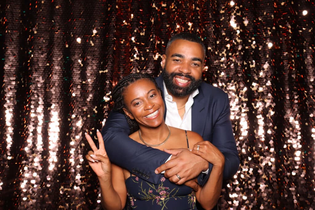 Couple posing playfully in a photo booth with a sparkling backdrop, woman smiling and making a peace sign, man embracing her from behind, capturing a joyful and intimate moment.