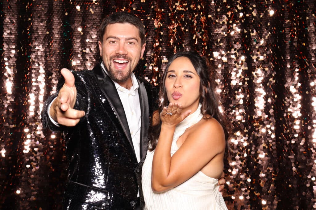 Couple posing playfully in a photo booth, man in a sequined jacket smiling and gesturing, woman blowing a kiss, shimmering backdrop enhancing the fun and romantic vibe.