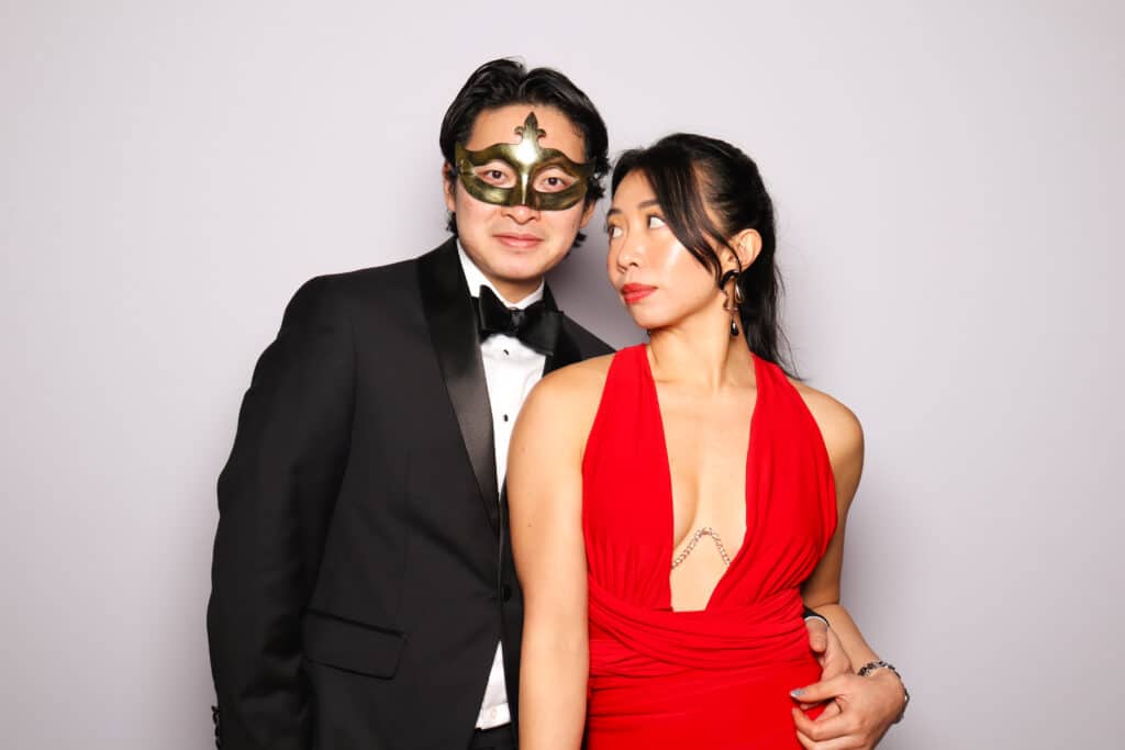 Couple posing in elegant attire, with a man in a tuxedo and green mask, and a woman in a red dress, exchanging playful glances, embodying flirtation and connection in a photo booth setting.