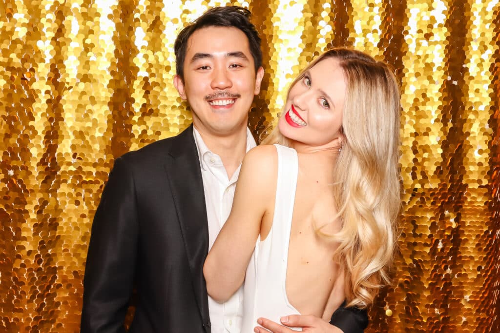 Couple posing playfully in a photo booth against a shimmering golden backdrop, capturing a joyful and intimate moment, embodying fun and romance.
