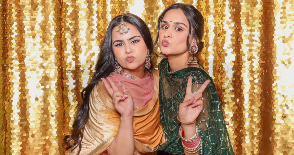 Two women posing playfully in traditional attire, making peace signs, with a sparkling golden backdrop, capturing a joyful moment at an event, reflecting Pop Life Photo's focus on candid interactions and real-time engagement in event photography.