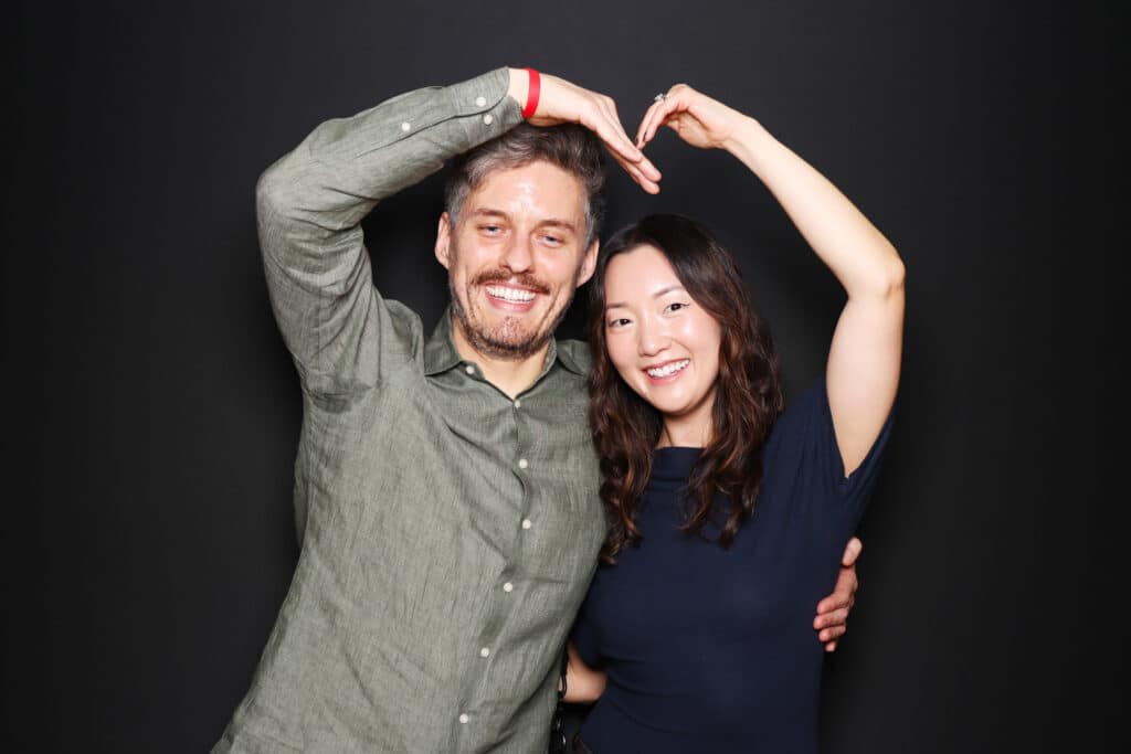 Couple creating a heart shape with their hands, smiling against a dark background, embodying playful and romantic photo booth pose for capturing genuine moments.