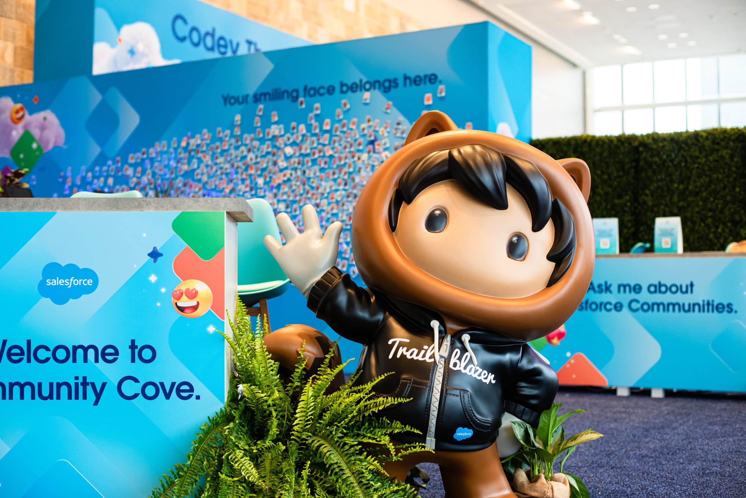 Trailblazer mascot welcoming attendees at Salesforce Community Cove, featuring vibrant blue backdrop with promotional text, plants, and event branding elements.