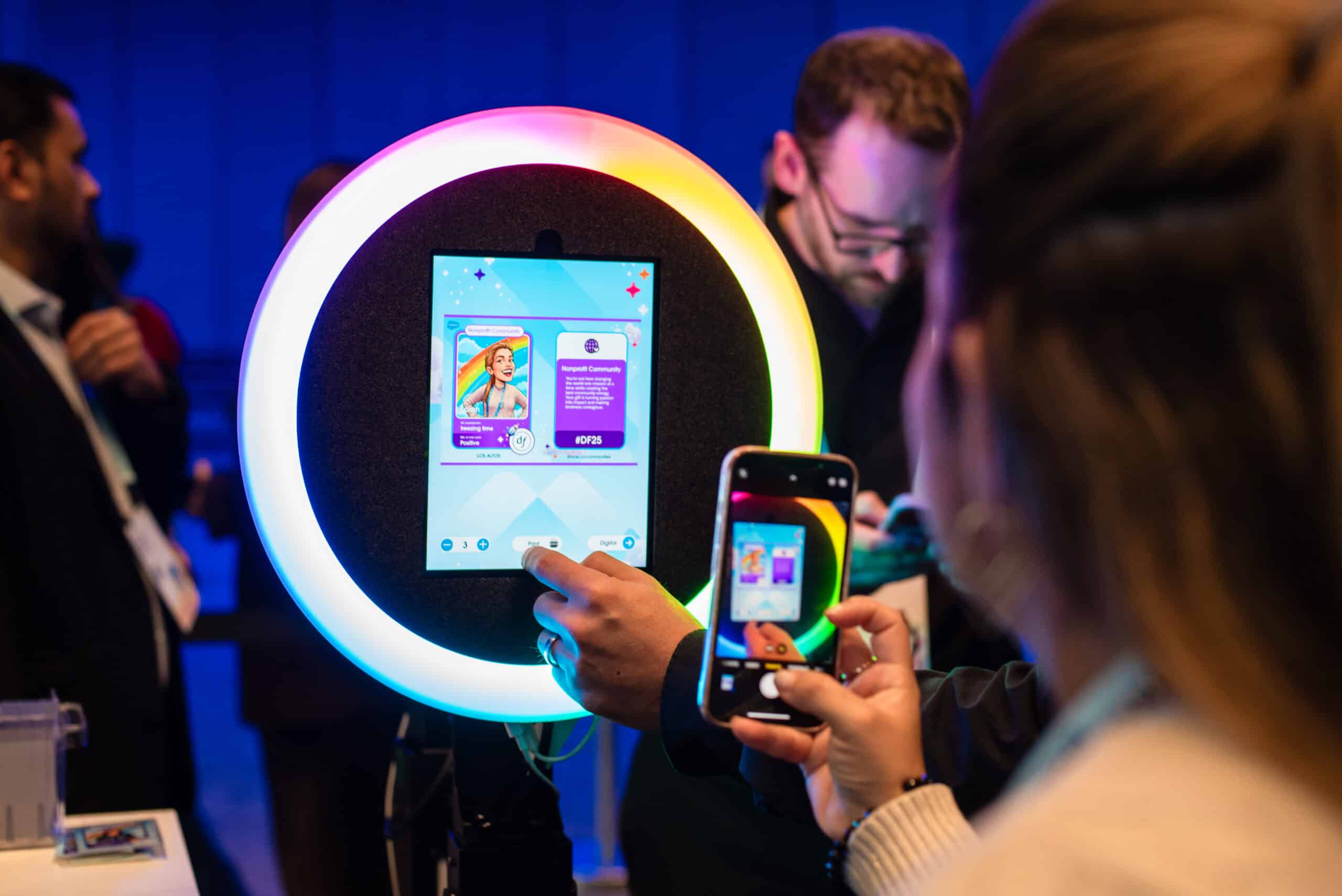 Person interacting with an AI-powered photo booth screen displaying colorful trading cards, with another attendee capturing the moment on a smartphone, at Dreamforce 2025 event.