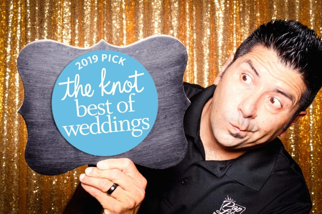 Man holding a sign that reads "2019 Pick the Knot Best of Weddings" in front of a sparkling gold backdrop, celebrating Pop Life Photo Booth's recognition in wedding services.