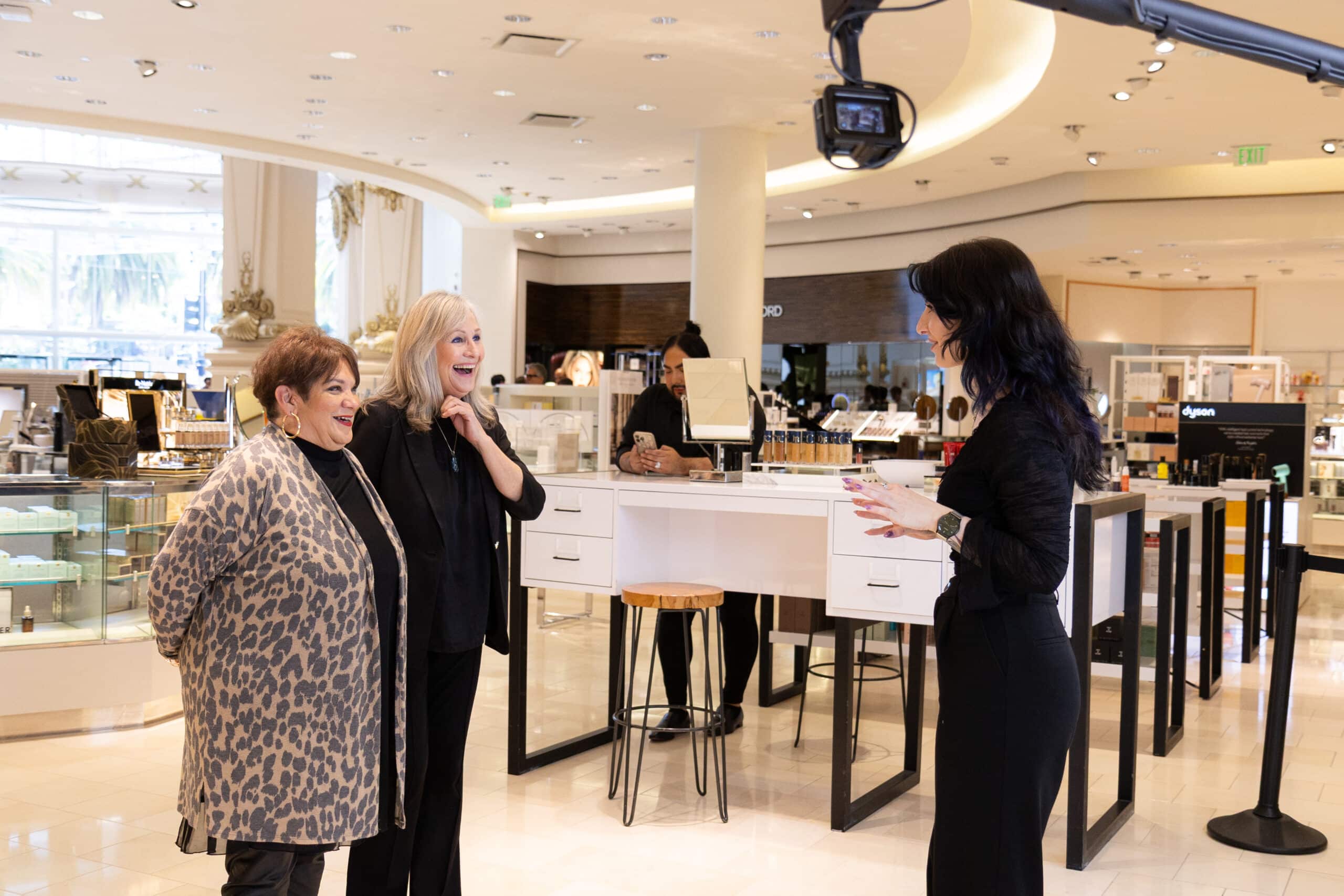 Two women smiling and interacting with a presenter in a luxury retail environment, showcasing the Glambot experience at Neiman Marcus, surrounded by beauty products and a camera setup for capturing shareable content.