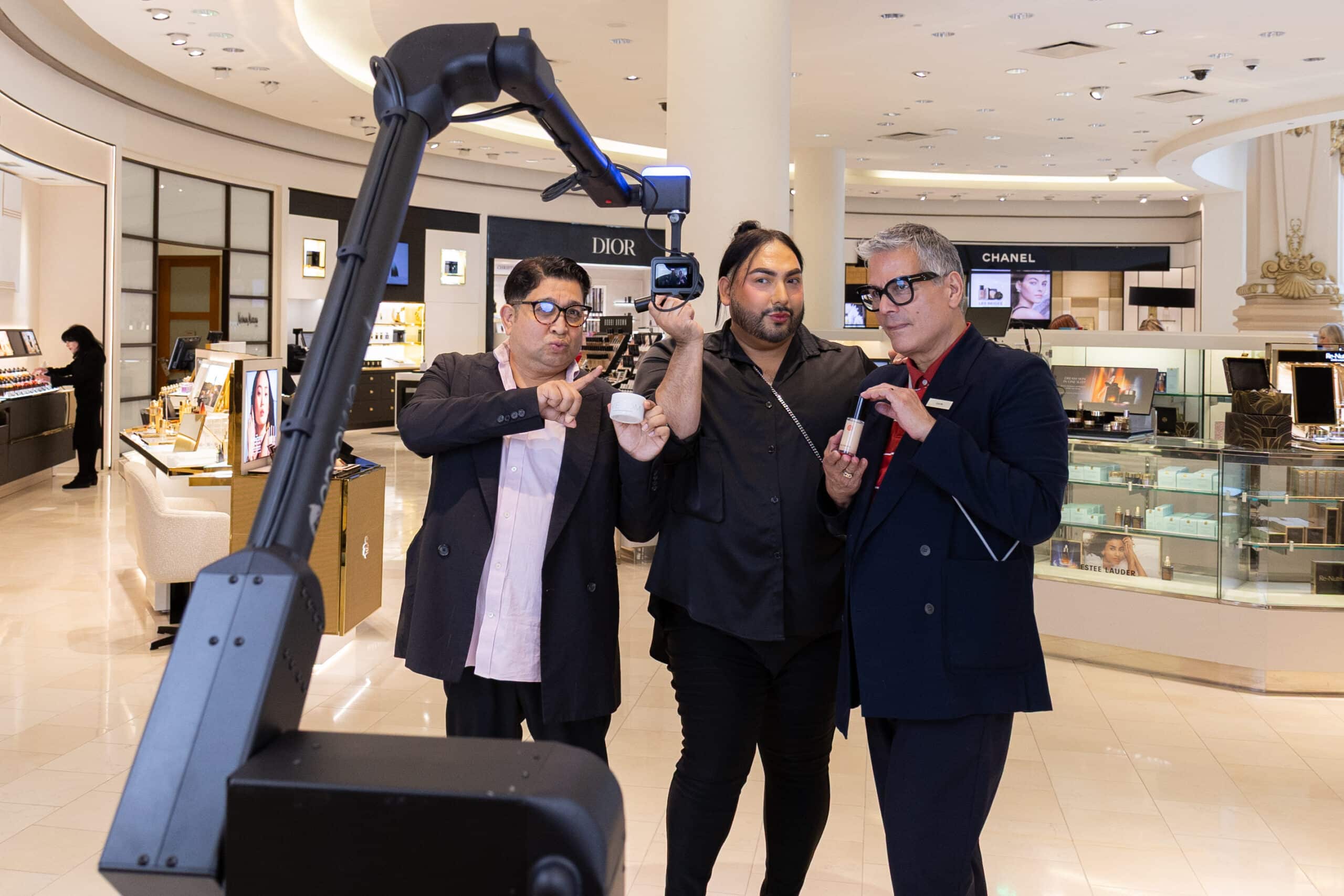 Three individuals showcasing beauty products in a luxury retail environment, featuring a Glambot camera setup, highlighting Neiman Marcus branding and a high-end shopping experience.