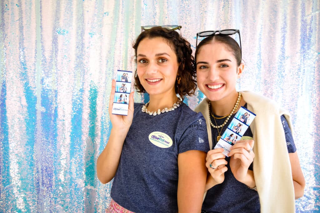 Two women smiling and holding photo booth strips in front of a shimmering backdrop, showcasing the Studio Booth experience for polished, editorial-style portraits at events.