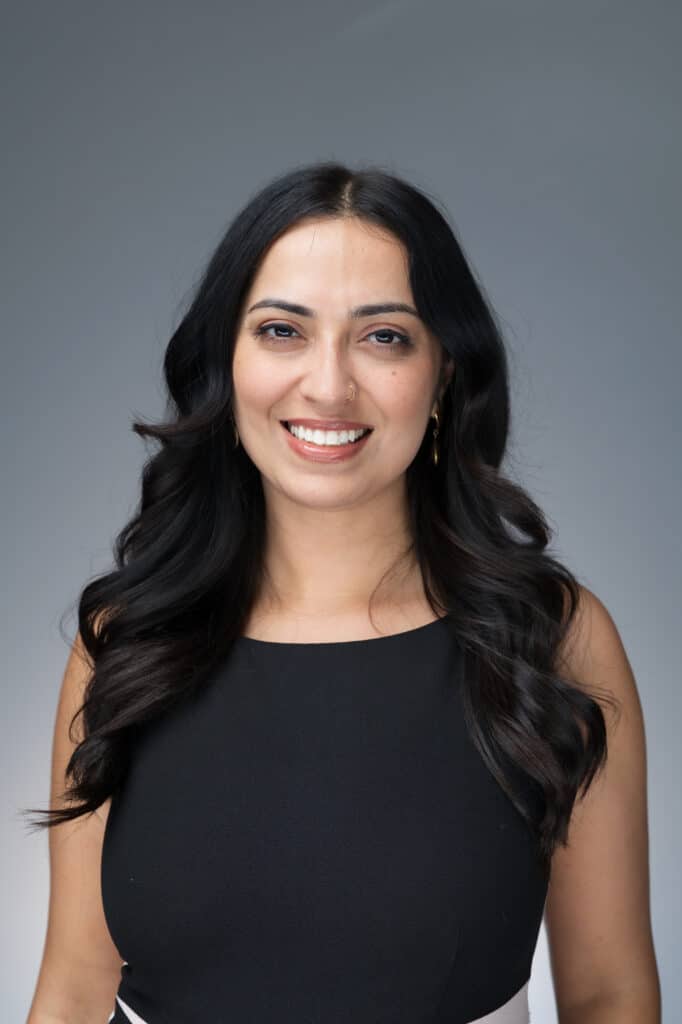 Professional headshot of a woman with long dark hair in a black top smiling against a neutral gray background.
