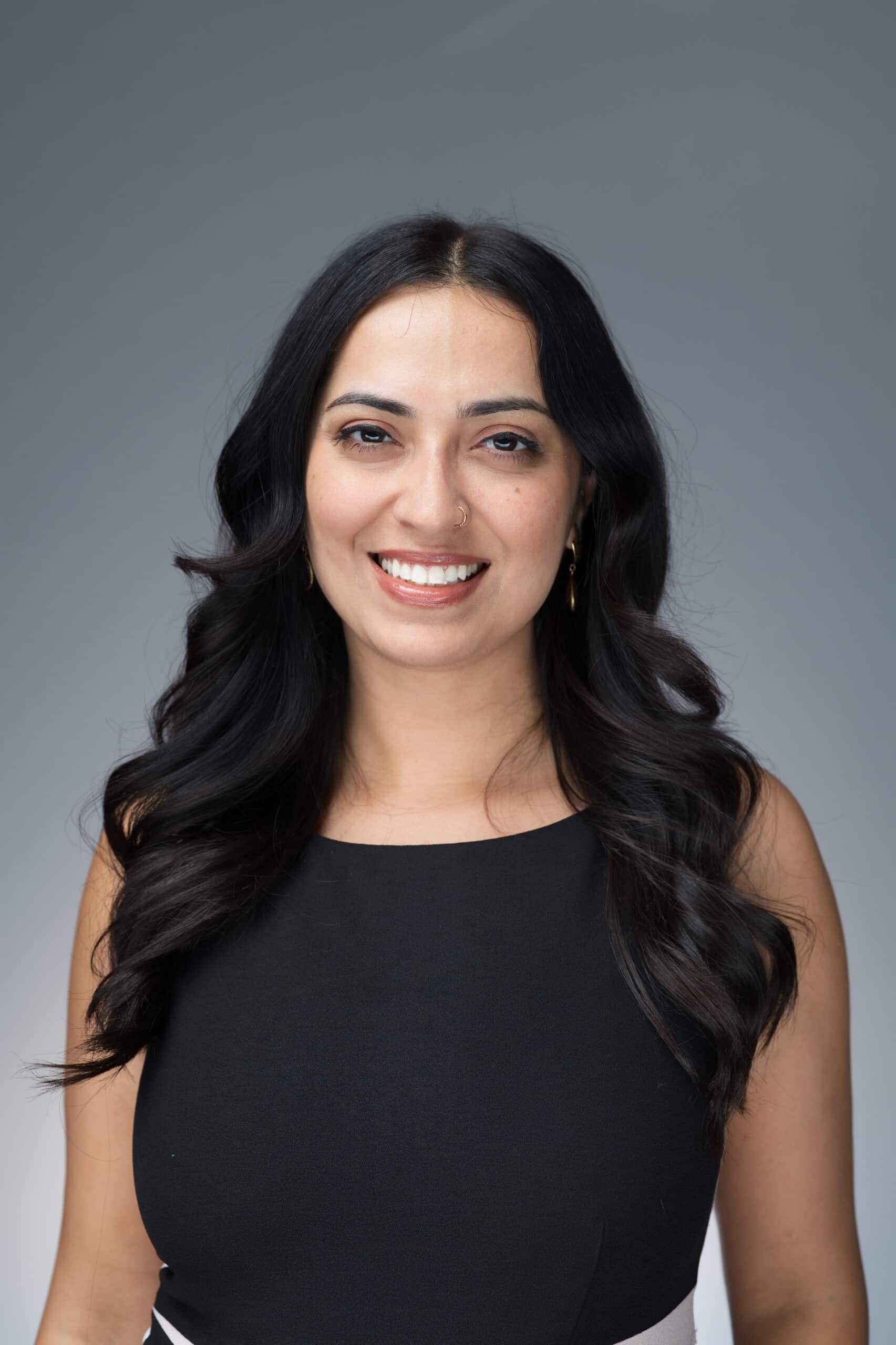 Professional headshot of a woman with long dark hair in a black top smiling against a neutral gray background.