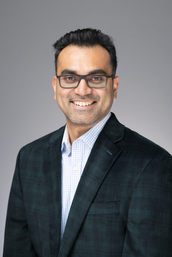 Professional headshot of a man in a dark blazer with a warm smile against a neutral gray background.