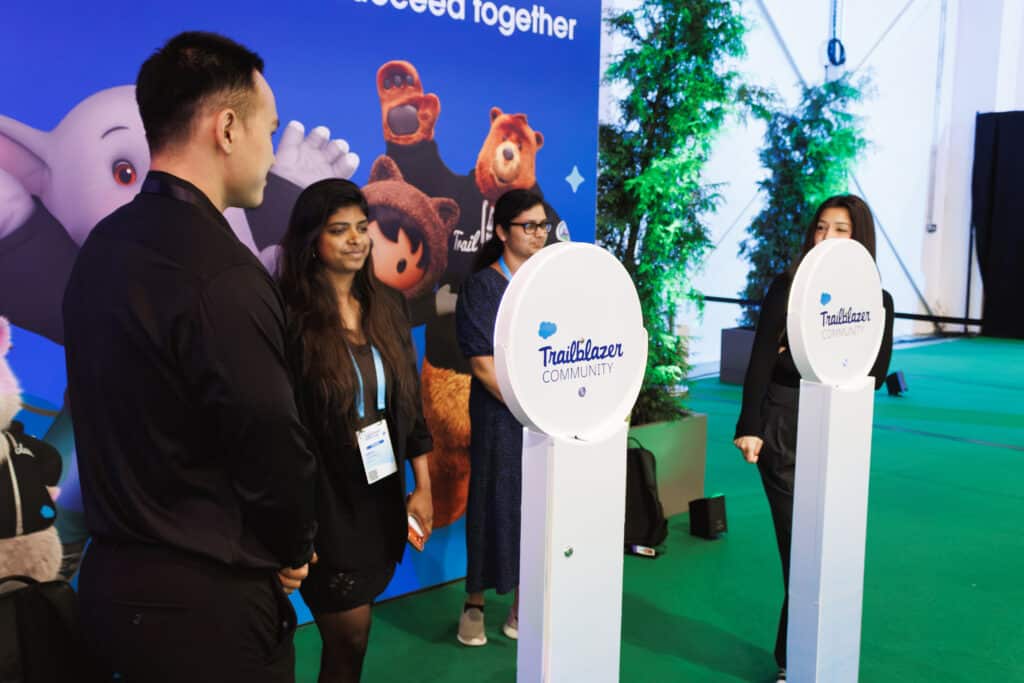 Attendees gathered at an event activation with two round "Trailblazer Community" photo stations, set against a blue backdrop featuring Salesforce cartoon mascots and the words "Succeed together," with green artificial turf and plants.