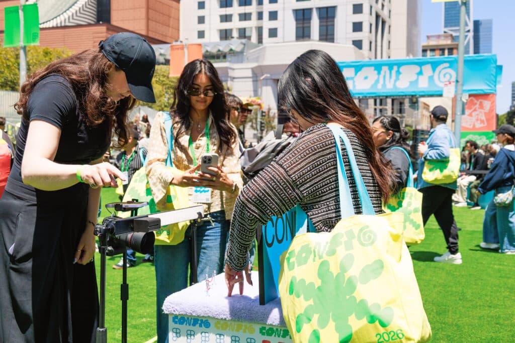 Attendees at an outdoor event on a city lawn, with women carrying green tote bags gathered around a display while one photographs it with a camera on a tripod, and colorful signage in the background.