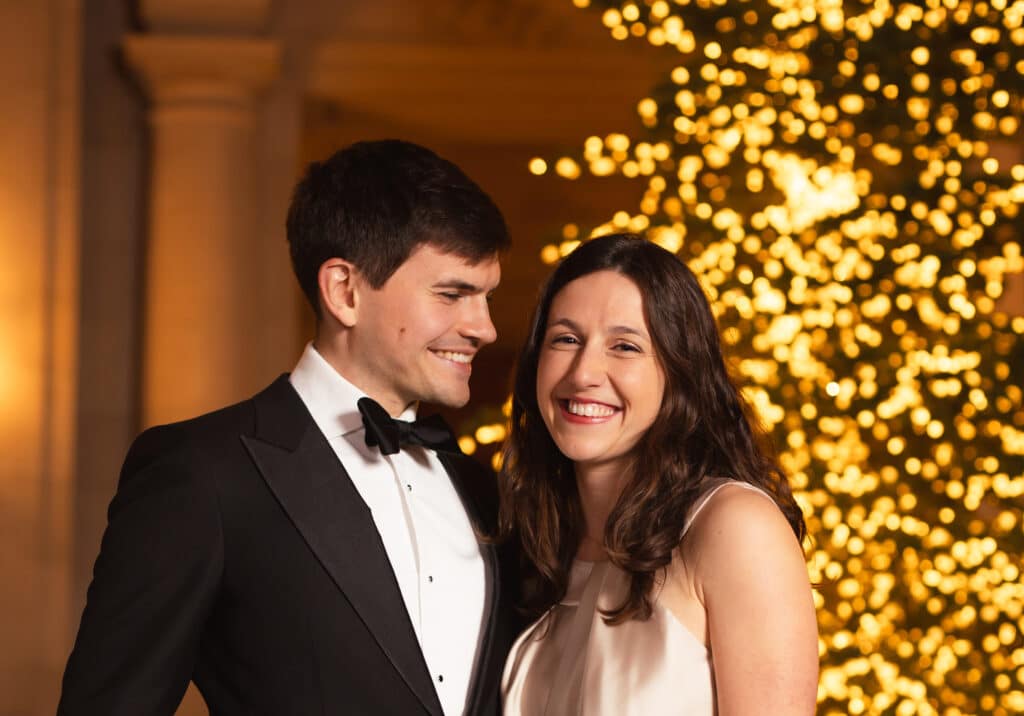 A smiling couple in formal evening wear at the McKinsey holiday celebration, a man in a black tuxedo and bow tie and a woman in a satin dress, posing in front of a tree covered in glowing golden lights.